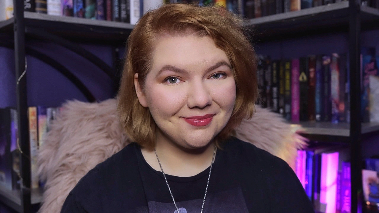 Jenna Streety Headshot. Purple and black bookshelf in background, pink fur cover on back of chair, black t-shirt on portrait of woman with short, caramel blond hair smiling at the camera.